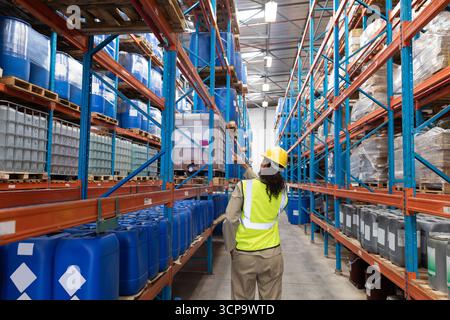Female warehouse worker wearing safety vest inspecting drums and pallets in warehouse aisle Stock Photo