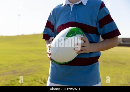 Teenage female athlete in striped rugby jersey holding white and green rugby ball on grassy field Stock Photo