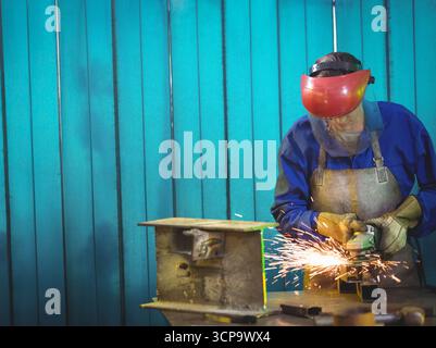 Female metalworker wearing face shield using angle grinder on shop bench with sparks, copy space Stock Photo