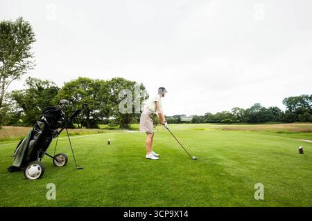 Senior female golfer holding driver while standing on tee box over golf ball and preparing drive Stock Photo