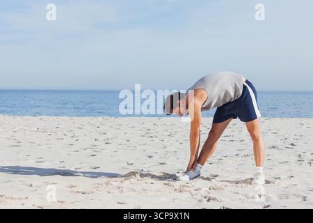 Man stretching forward in sleeveless top, sports shorts, athletic shoes on sandy beach, copy space Stock Photo