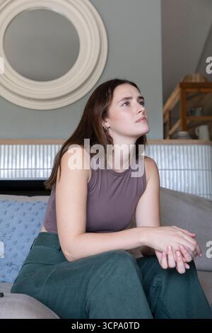 Woman sitting on sofa with throw pillow in living room gazing toward round mirror and shelf Stock Photo