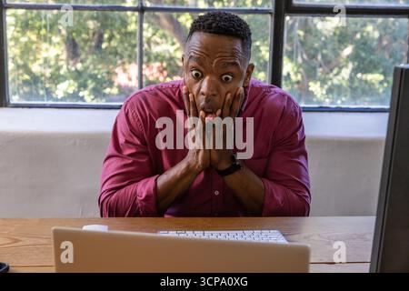 African american man pressing cheeks and showing shock at home desk with laptop Stock Photo