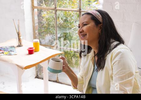 Middle-aged Hispanic woman smiling while holding ceramic mug beside painting supplies in art studio Stock Photo
