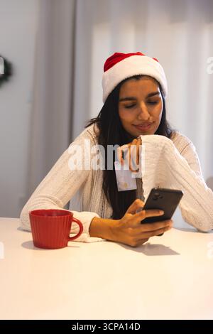 Woman wearing Santa sweater and hat, holding credit card, using smartphone at table with mug Stock Photo