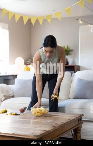 Asian woman arranging glass bowl with chips, party blowers on coffee table in living room Stock Photo