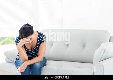 Mature woman leaning forward on gray sofa in living room under white blinds wearing T-shirt, jeans Stock Photo