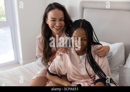 Diverse female friends hugging and laughing on bed in bedroom near window wearing pink silk robes Stock Photo