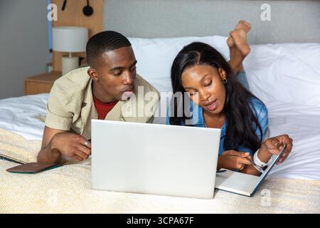 African American couple lying on bed at home pointing at open notebook while watching laptop screen Stock Photo
