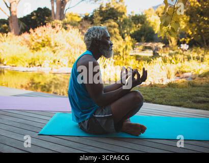 Senior African American man practicing meditation on turquoise yoga mat on wooden deck beside pond Stock Photo
