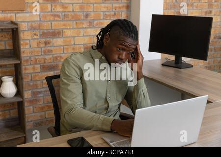 African american man sitting at desk in brick office working on laptop with smartphone Stock Photo