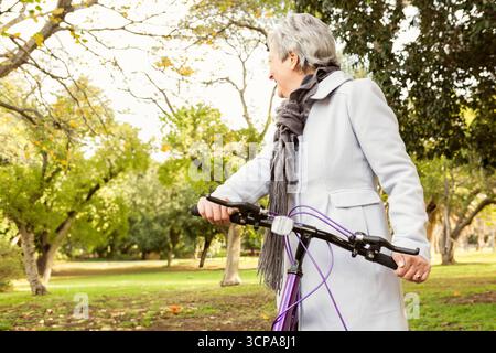 Senior woman holding bicycle and wearing long coat with scarf in leafy autumn park, copy space Stock Photo
