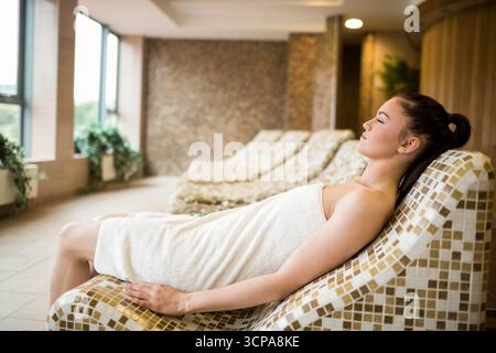 Woman wearing towel reclining on mosaic-tiled lounge chair at spa with large windows, copy space Stock Photo