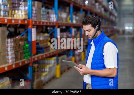 Warehouse worker checking inventory on tablet and wearing headset in aisle lined with boxes Stock Photo