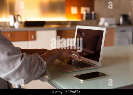 Man typing on laptop at kitchen workspace showing smartphone, kettle and coffee maker, copy space Stock Photo