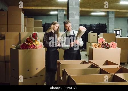 Two floral workers in a warehouse are discussing arrangements while surrounded by boxes and colorful flower bouquets, showcasing teamwork and Stock Photo