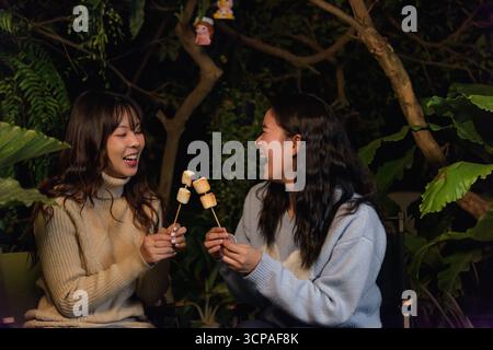 Winter camp outdoor, two happy women friends enjoying forest evening, roasting delicious marshmallow Stock Photo