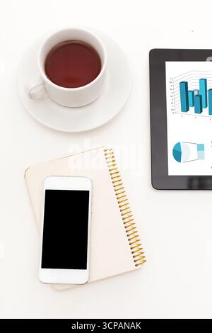 Busy workspace with coffee, smartphone, notebook, and tablet displaying data graphs Stock Photo
