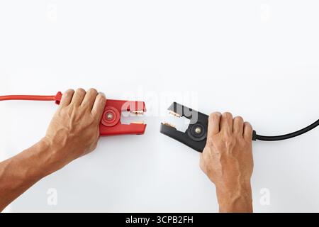 Two hands holding car jumper cable clamps, one red and one black, facing each other on a plain white background Stock Photo