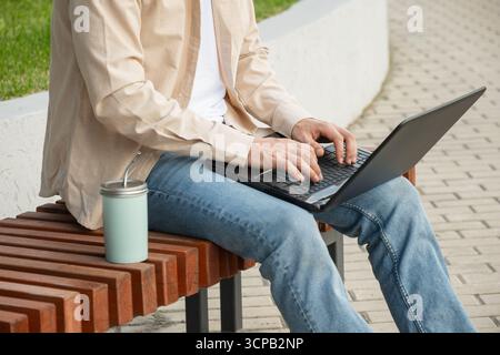 Handsome man, attractive freelancer , brunette sits on bench in urban park, using laptop for remote work. Wearing casual clothes. Outdoors. Stock Photo