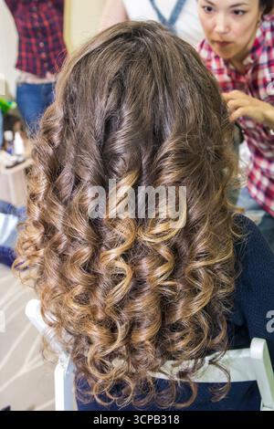 A woman with long, curly hair is getting her hair done by another woman. The scene is set in a salon, with a chair and a table in the background Stock Photo