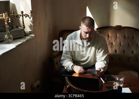 Caucasian young adult man sitting on vintage sofa reading document with focused expression, smartphone and glass of water on table, sunlight streaming through window illuminating scene Stock Photo