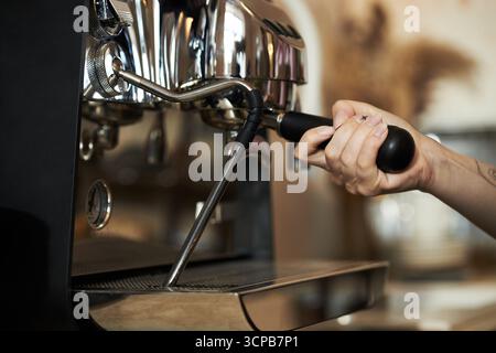 Caucasian young adult woman operating espresso machine, gripping portafilter handle with right hand, preparing coffee in cafe setting, visible tattoo on forearm, closeup view Stock Photo