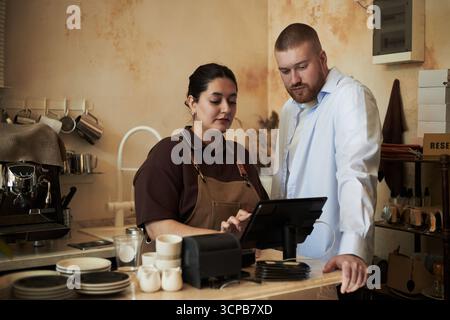 Caucasian young adult man and Hispanic young adult woman standing at counter using touchscreen point of sale terminal in coffee shop, both focusing on digital transaction process Stock Photo