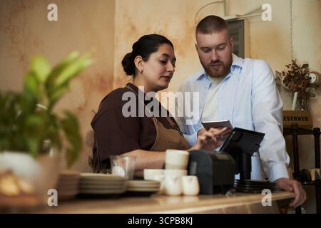 Caucasian young adult man and Hispanic young adult woman standing behind counter using touchscreen point of sale terminal, both focused on screen while discussing transaction Stock Photo