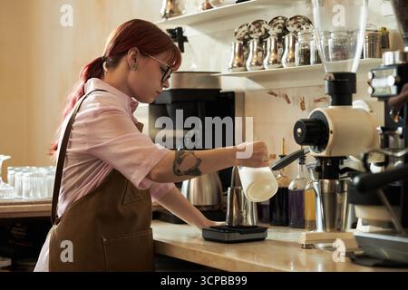 Young adult Caucasian woman with red hair preparing coffee using milk frother and scale in cafe, wearing apron and glasses, standing at counter with professional coffee equipment Stock Photo
