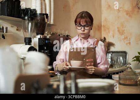 Young adult Caucasian woman wearing glasses standing behind counter holding cup and saucer in coffee shop, looking down with neutral expression, surrounded by coffee equipment Stock Photo