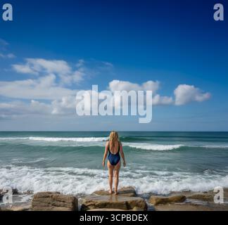 A woman in a swimsuit stands on rocks, contemplating the ocean waves before a refreshing wild swim on a beautiful sunny day. Stock Photo