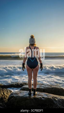 A woman in a bobble hat and gloves stands on rocks, ready for a cold water sea swim as the sun rises over the waves. Stock Photo