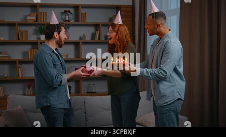 Three multiracial friends at home party birthday congratulation couple Caucasian woman with African American male guy congratulate man giving present Stock Photo