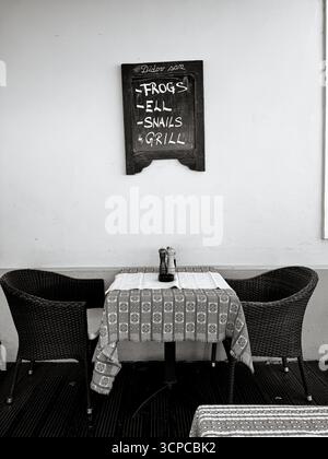 A minimalist black-and-white view of a Zagreb café table set for two, beneath a chalkboard menu advertising “Frogs, Eel, Snails, Grill,” Stock Photo