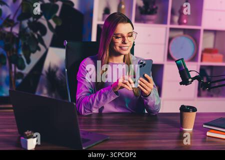 Young woman in a modern studio setup engaging with her phone and surrounded by multimedia tools for content creation Stock Photo