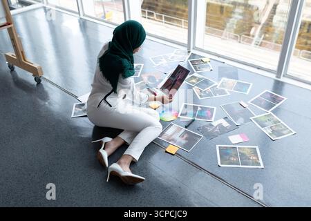Middle Eastern hijabi woman sitting on studio floor, reviewing prints, color wheel and sticky notes Stock Photo