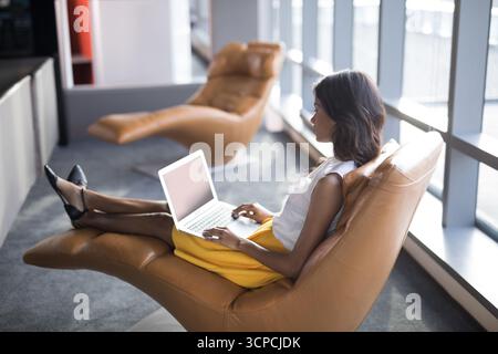 Asian African American woman reclining on chaise lounge at office with laptop, copy space Stock Photo