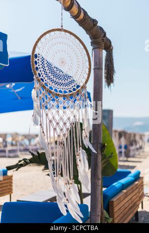 A beautiful dreamcatcher is hanging from a wooden pole, gently swaying in the breeze. Bright blue umbrellas shade nearby lounge seating on a sandy bea Stock Photo
