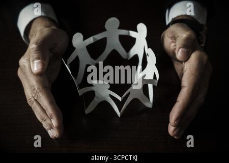 Businessman Hands Protecting a Circle of Paper Chain People on Dark Wooden Table Stock Photo