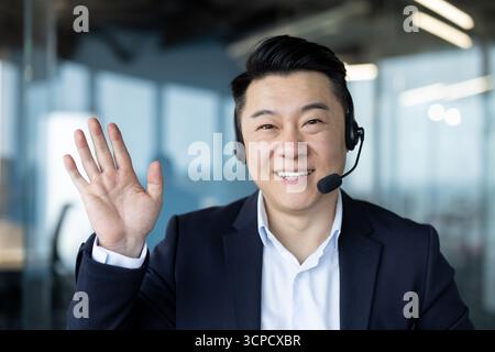 Close-up portrait of a smiling young Asian man in a suit and headset sitting in an office facing the camera, greeting and waving. Stock Photo