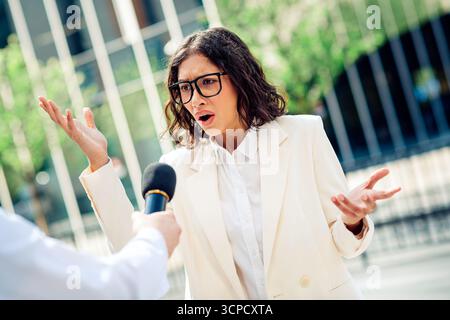 Confident young businesswoman answering questions during an outdoor business interview near a modern urban office building Stock Photo