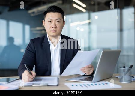 An asian businessman working in an office setting. He is dressed in a suit and working on documents at his desk. Focus is on him. Stock Photo