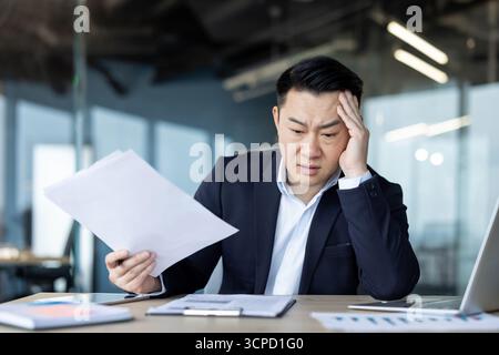An asian businessman looks stressed while reviewing documents at his desk. He is wearing a suit and working in an office environment. Stock Photo