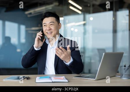 Smiling young Asian businessman in suit sitting in office at desk and talking while gesturing with hands. Stock Photo