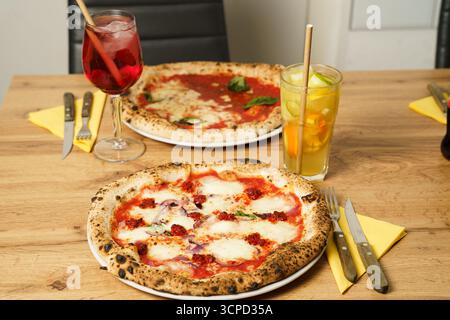 Two freshly baked Neapolitan-style pizzas served on a rustic wooden table, accompanied by colorful cocktails—an Aperol Spritz and a citrus-infused dri Stock Photo