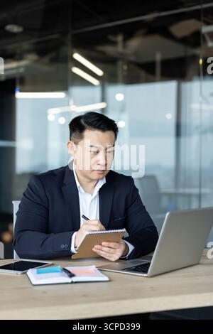 An asian businessman sits at his desk, concentrating as he writes notes and works on his laptop in the office. Stock Photo