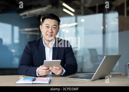 Portrait of a smiling young man, Asian businessman and office worker sitting at a desk, holding a tablet and looking at the camera. Stock Photo