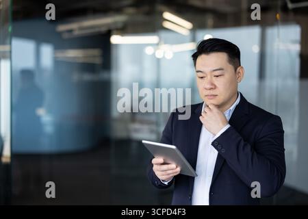 A thoughtful young Asian man is standing inside an office and looking seriously at the screen of a tablet he is holding in his hand. Stock Photo
