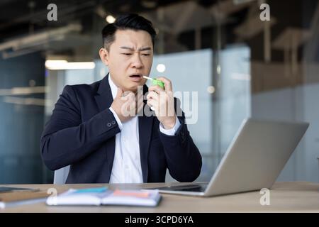Asian young man sitting in the office at his workplace and using an aerosol for asthma and allergies. Stock Photo